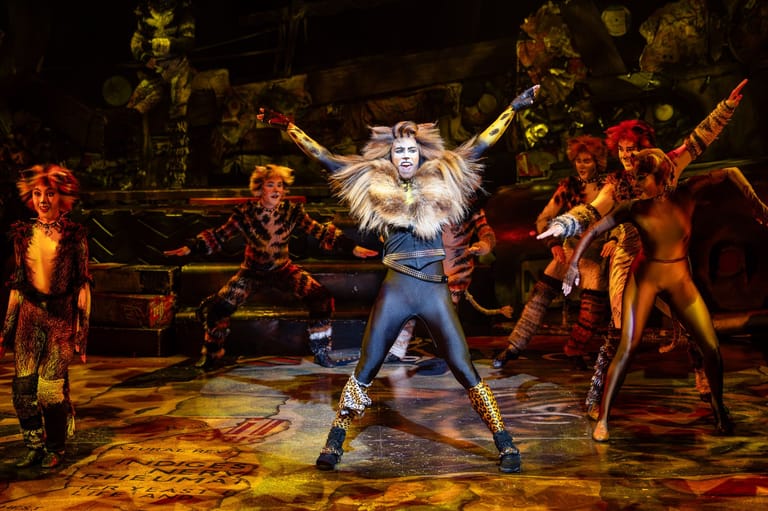 Performer in white hair and blue outfit strikes a dynamic pose centre stage, surrounded by dancers in animal costumes under warm theatrical lighting.