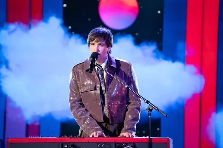 Musician performing at a keyboard with microphone against a colourful stage backdrop of clouds and neon lighting.
