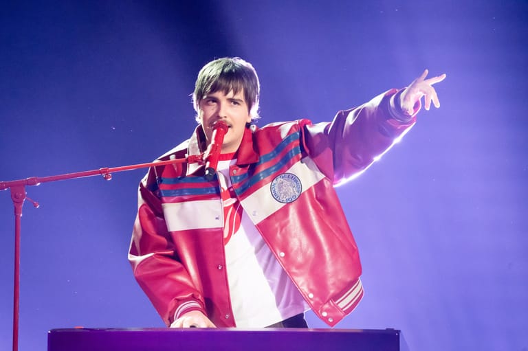 Performer in red and white jacket singing at microphone with arm extended, purple stage lighting behind.