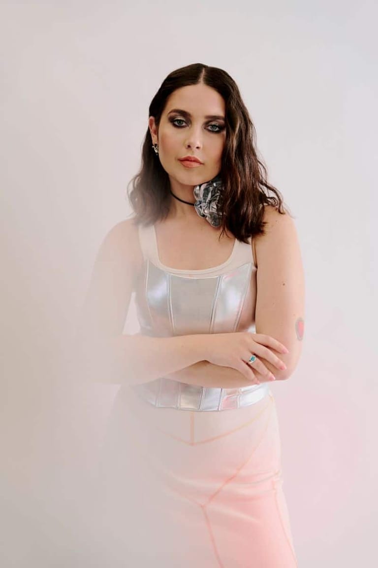 Woman with dark shoulder-length hair wearing a white sleeveless top and pink skirt against a neutral background.