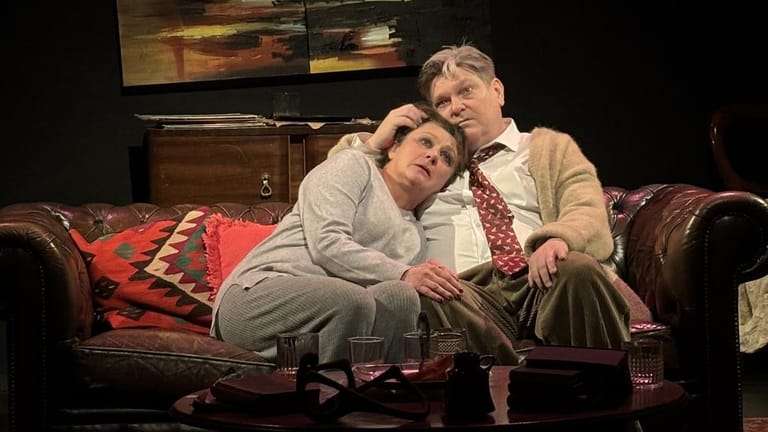 Two performers seated on a red patterned sofa during a theatrical scene, one resting against the other's shoulder.