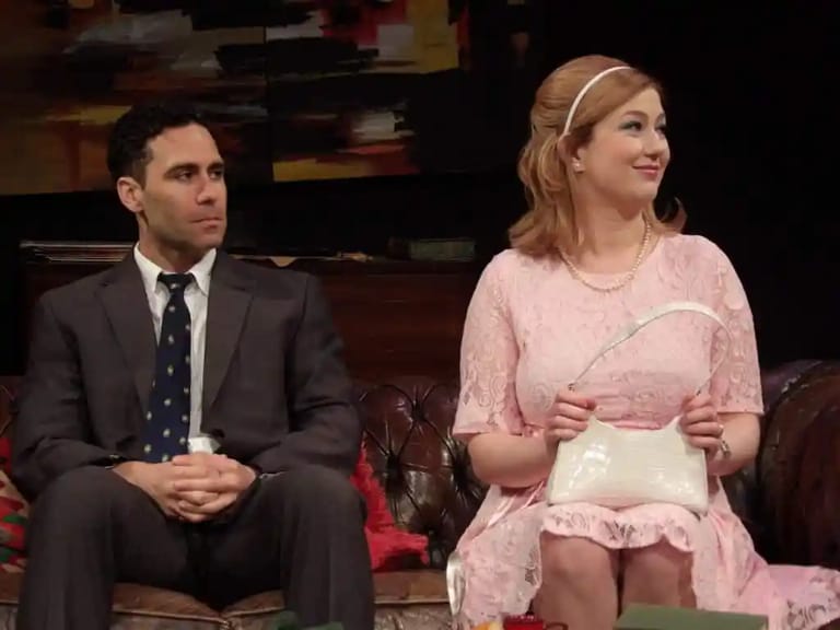 Two people sit on a red sofa during an interview, the man in a dark suit and the woman in a pink dress with a headband.