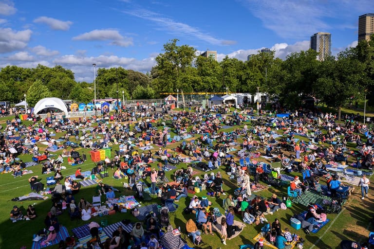 Large outdoor crowd gathered on a grass field under blue sky with trees and city buildings visible in the background.