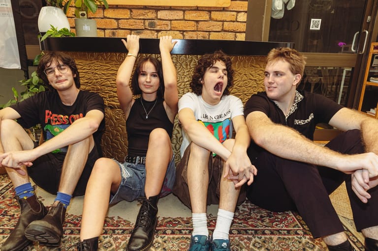 Four young musicians sit casually against a brick wall, laughing and relaxed in a candid band portrait.