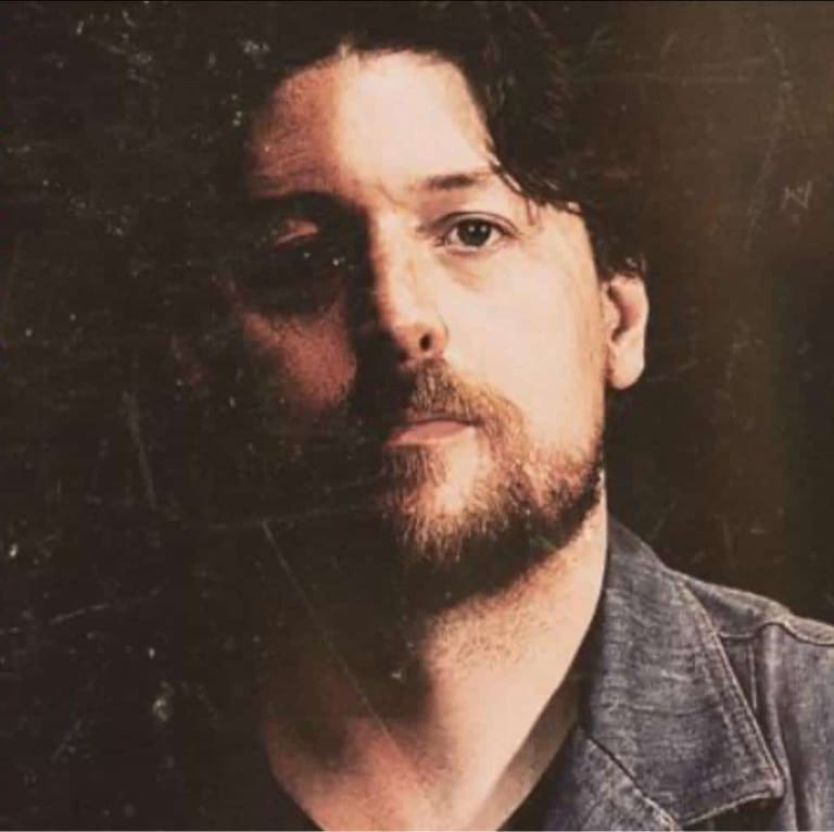 Man with dark hair and beard in denim shirt against dark background, looking directly at camera.