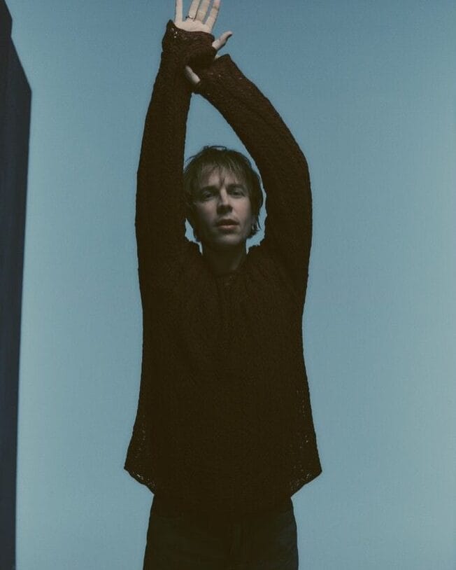Person in black suit with arms raised overhead against pale blue background.