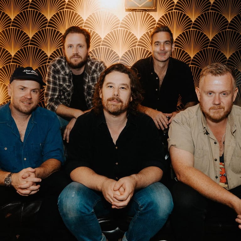 Five men pose together against a gold art deco patterned backdrop during an interview setting.