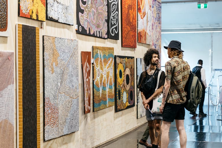 Gallery visitors examine Indigenous artworks displayed on cream walls in a bright, modern exhibition space.