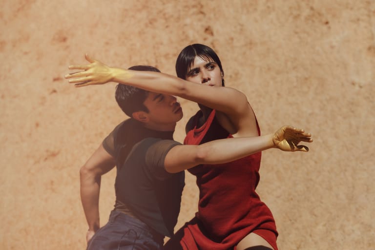 Two dancers in close contact against a textured neutral wall, arms extended in a contemporary dance pose.