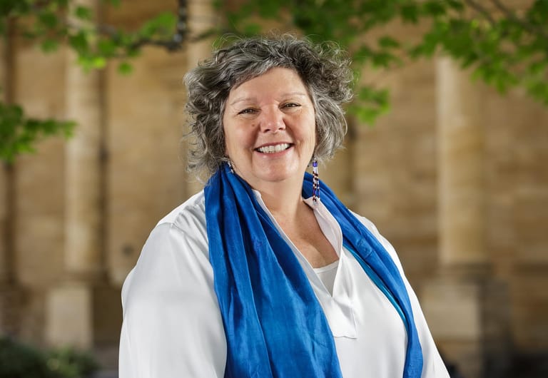 Woman with grey curly hair wearing white shirt and blue scarf, smiling at camera against stone wall.