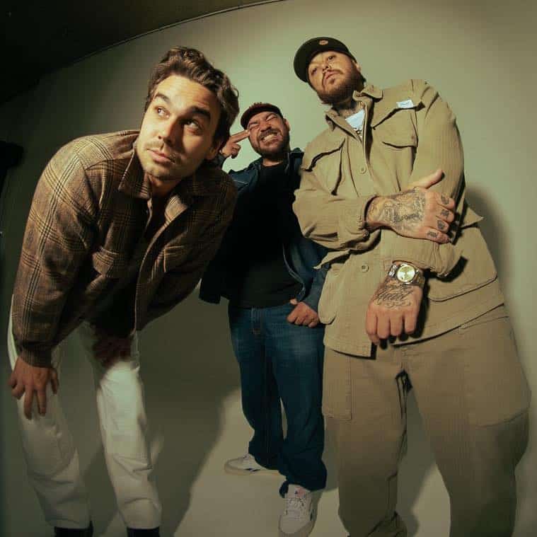 Three men pose together against a neutral backdrop, with one leaning forward and two standing beside a large cardboard cutout figure.