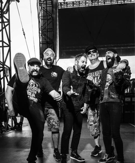 Five musicians in dark clothing and caps pose energetically on stage, laughing and gesturing during an interview.