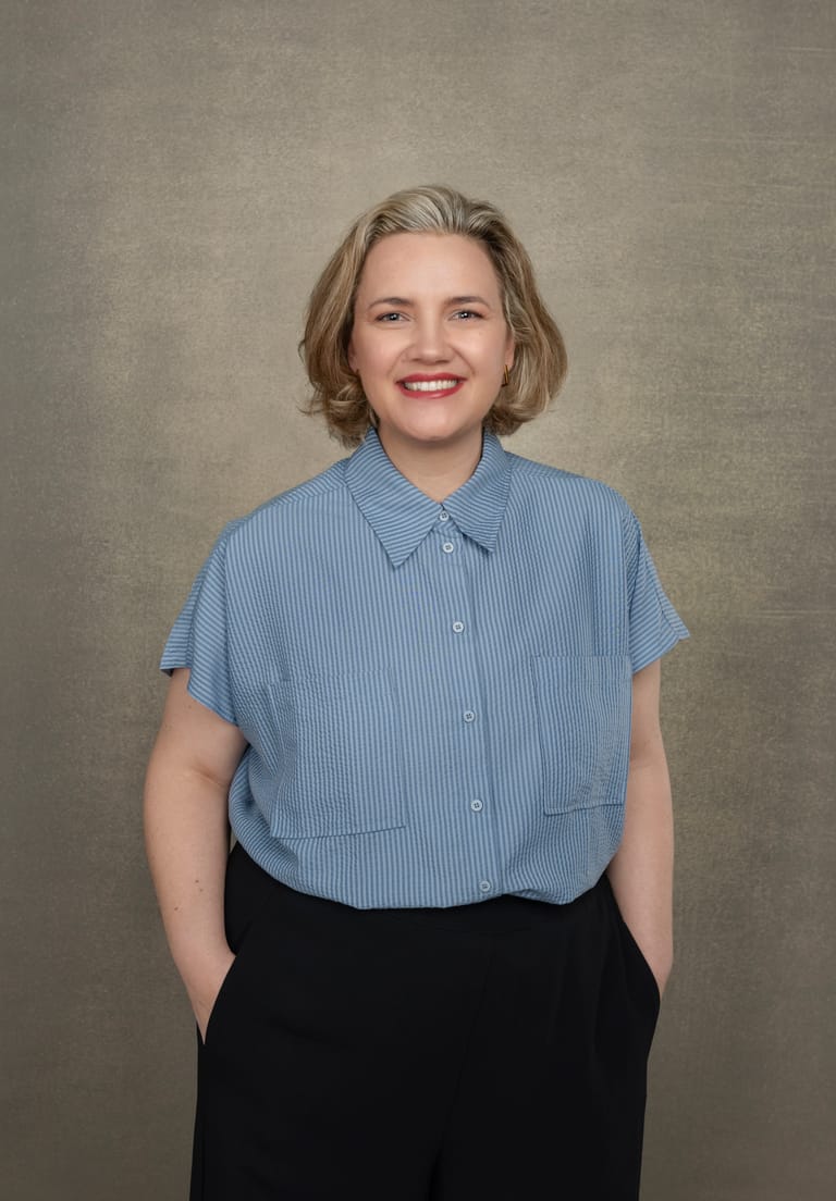 Woman with shoulder-length blonde hair wearing a light blue striped shirt and black trousers, smiling against a neutral beige background.