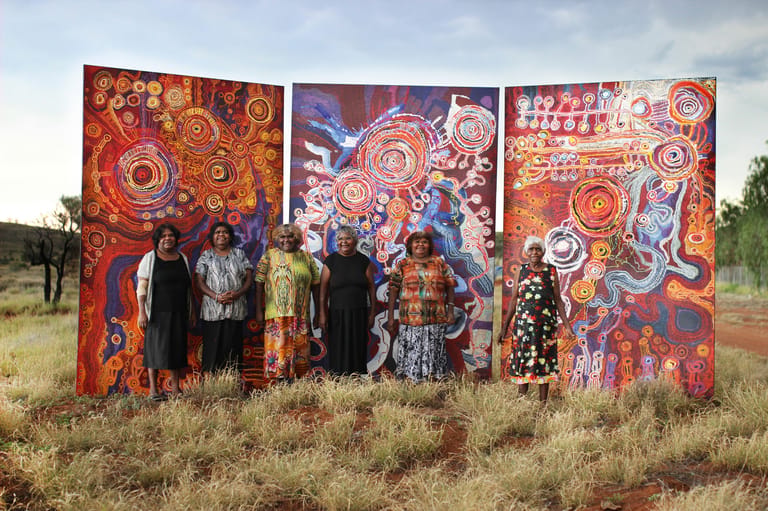 Group of people standing before three large colourful Indigenous artworks displaying circular patterns in an outback setting.