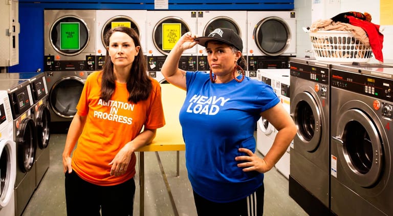 Two people pose confidently in a laundry facility surrounded by washing machines and dryers.