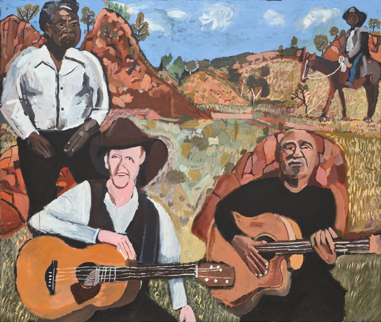 Two guitarists perform outdoors against a painted backdrop of red rock formations and desert landscape with figures on horseback.