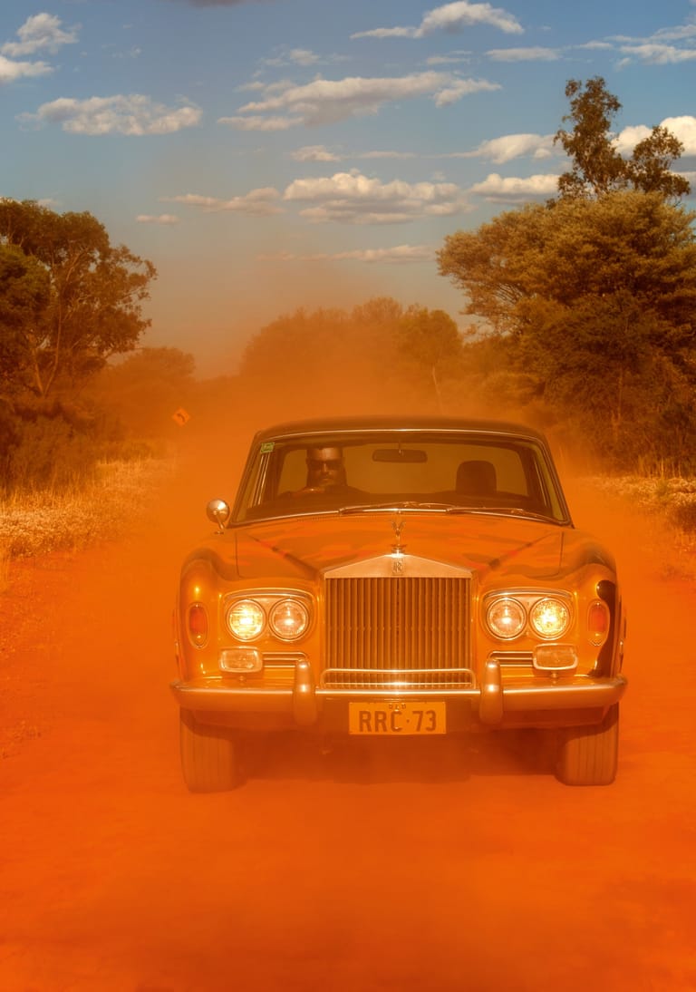 Vintage Rolls-Royce driving through a dusty orange outback landscape with sparse eucalyptus trees.