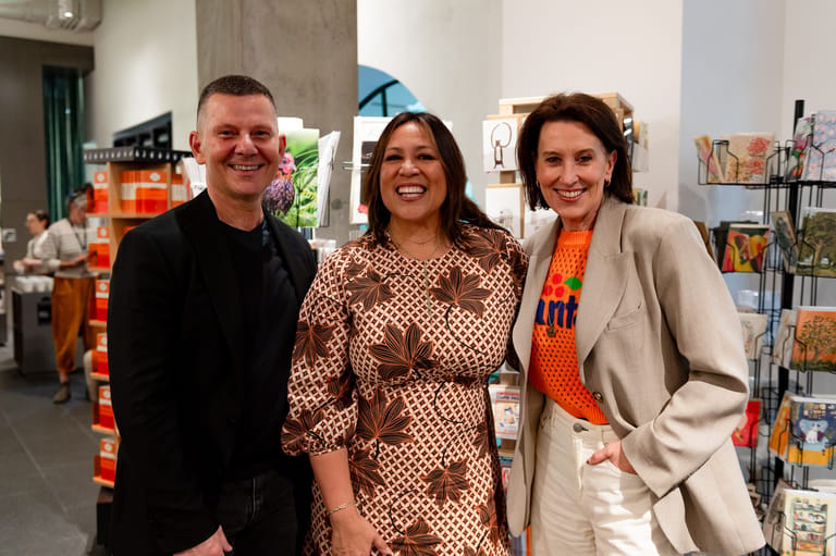 Three people stand smiling in a colourful bookstore or arts venue, with shelves and displays visible behind them.