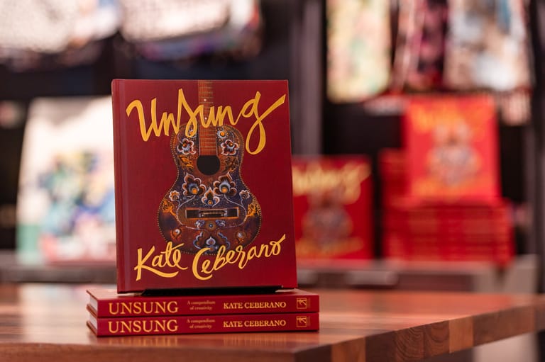 Stack of red "Unsung" books by Kate Ceberano displayed on a wooden surface with blurred background items.