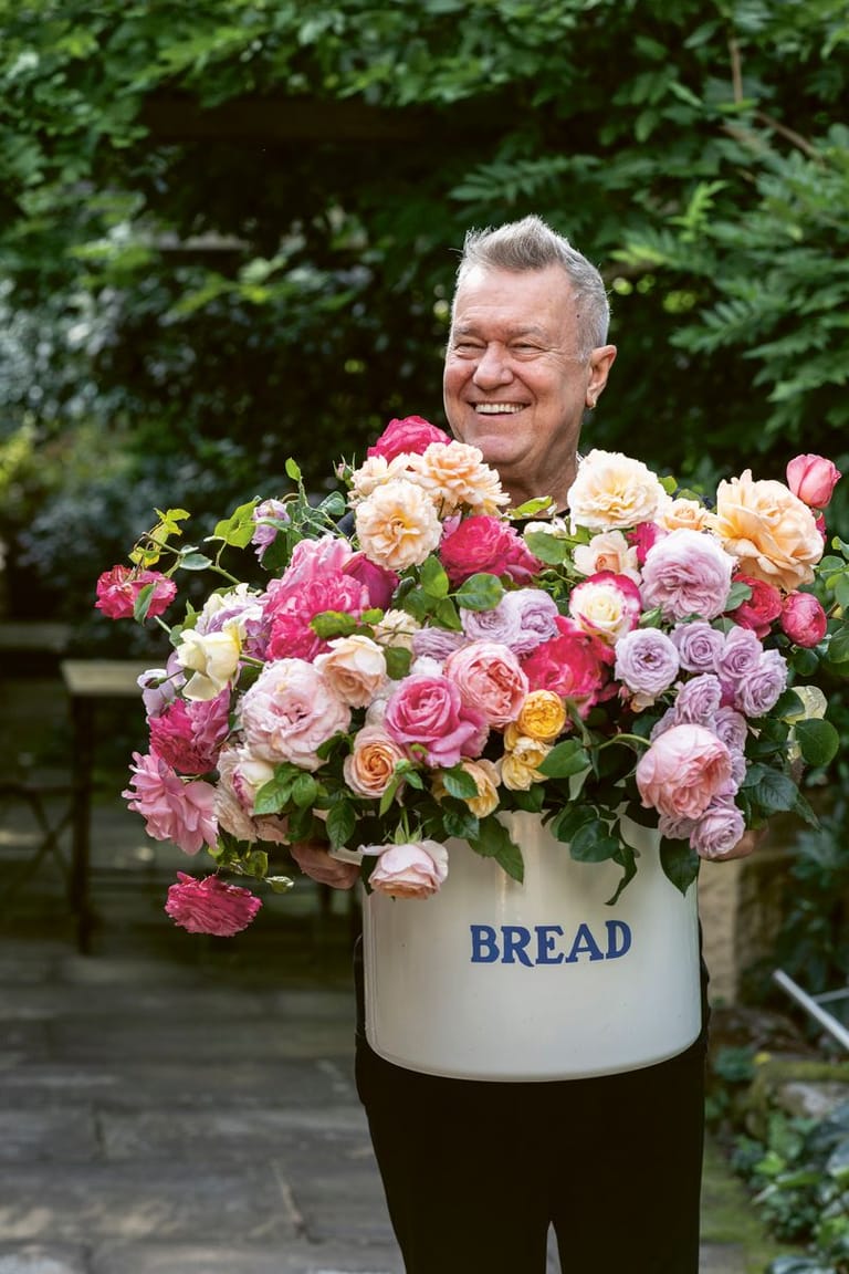 Man smiling behind an overflowing arrangement of pink, cream and yellow roses in a vintage bread crock.