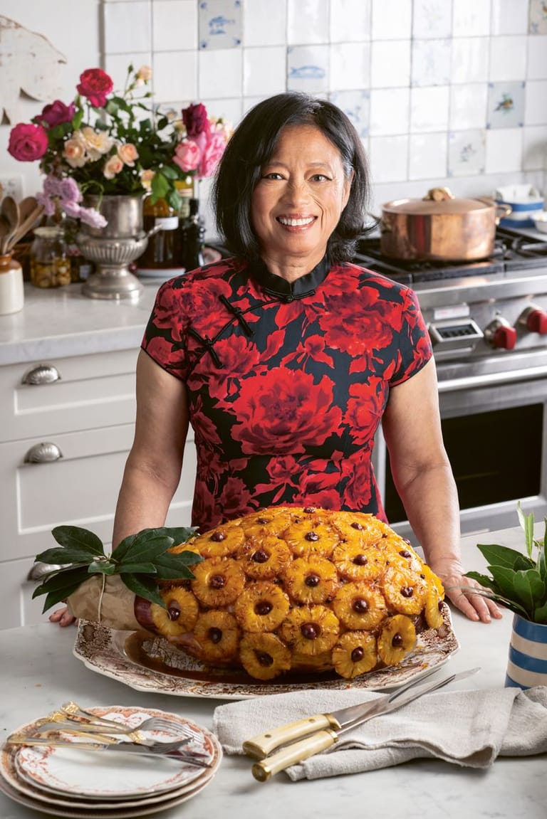 Woman in a red floral shirt stands behind a golden ring cake in a bright kitchen with fresh flowers.