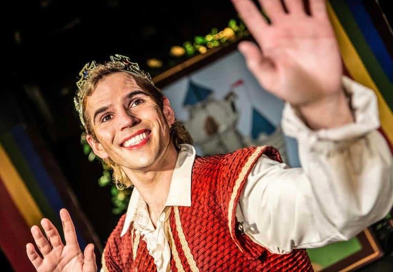 Performer in red embroidered waistcoat and white shirt waves to audience under warm stage lighting.