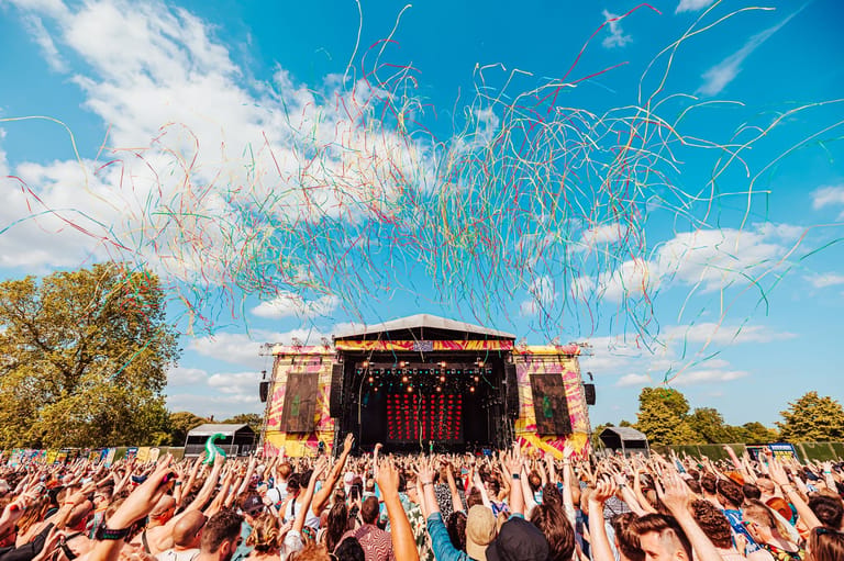 Crowd of festival-goers with raised arms facing a stage beneath blue sky and decorative light installation.