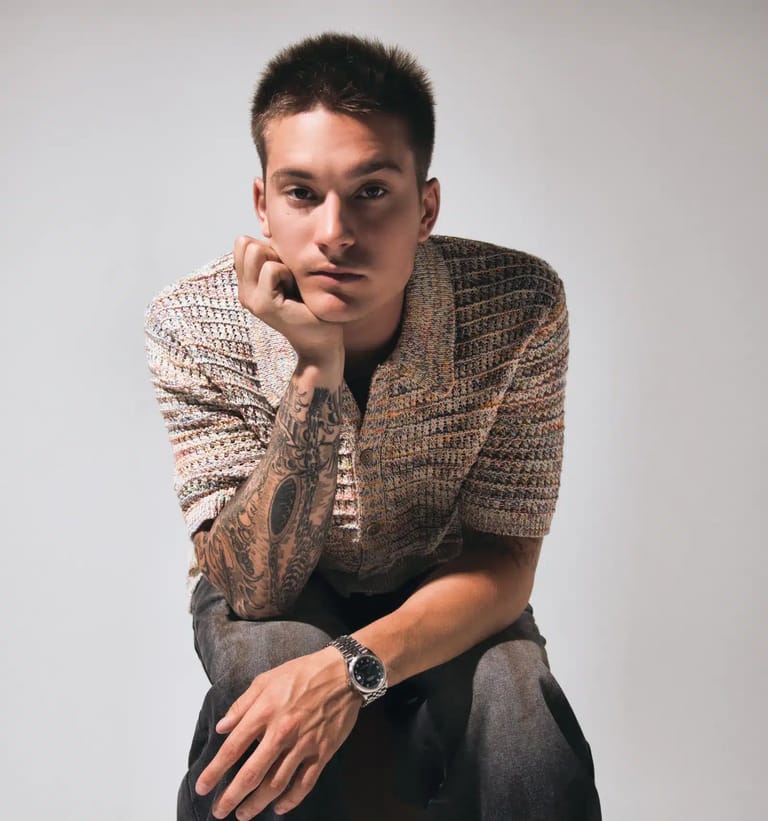 Young man in a textured shirt with tattooed arm, resting chin on hand against a neutral background.