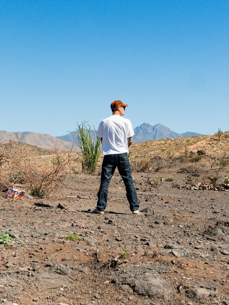Person standing on arid terrain with mountain range visible under clear blue sky.