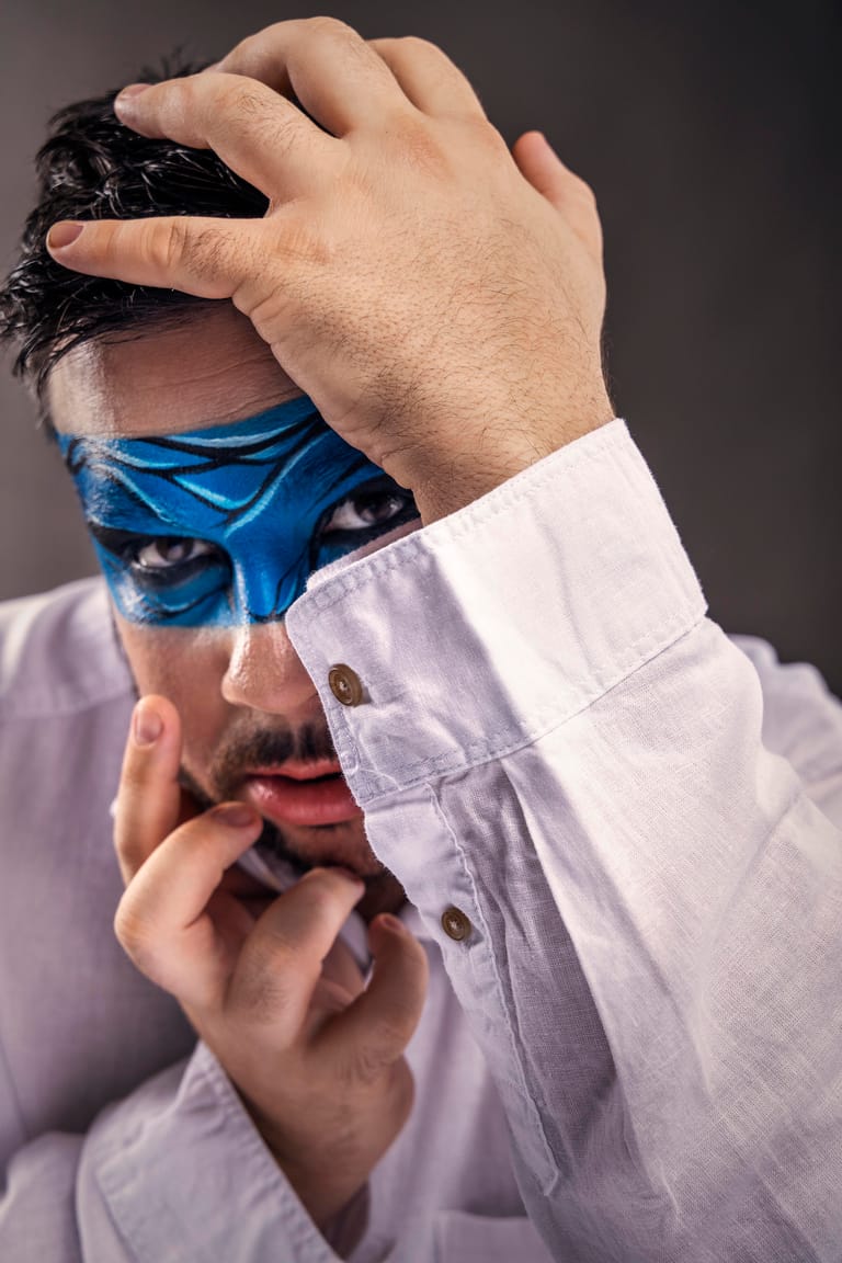 Person in white shirt with bright blue face paint removes a black mask against grey background.