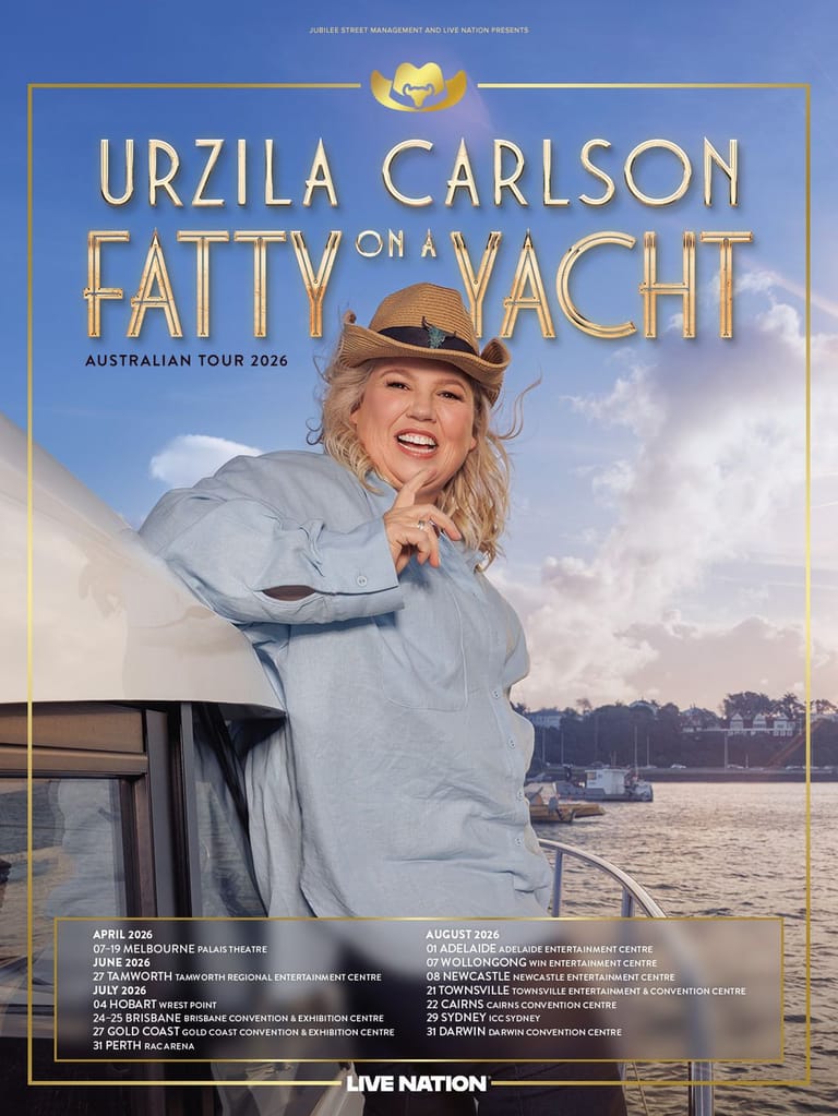 Performer in a light blue jacket and brown hat smiling on a yacht, with water and sky visible behind her.