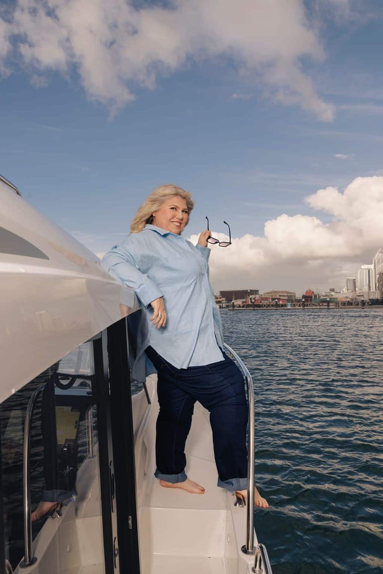 Woman in light blue shirt standing on the side of a boat on blue water with a city skyline in the background.