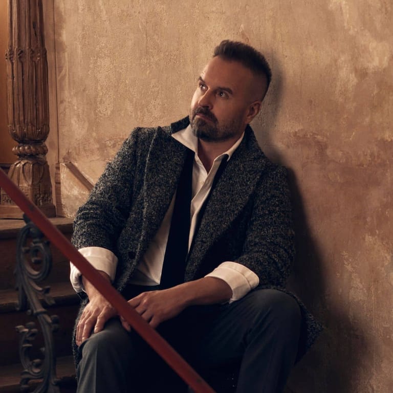 Bearded man in dark tweed jacket and white shirt seated against textured gold wall.
