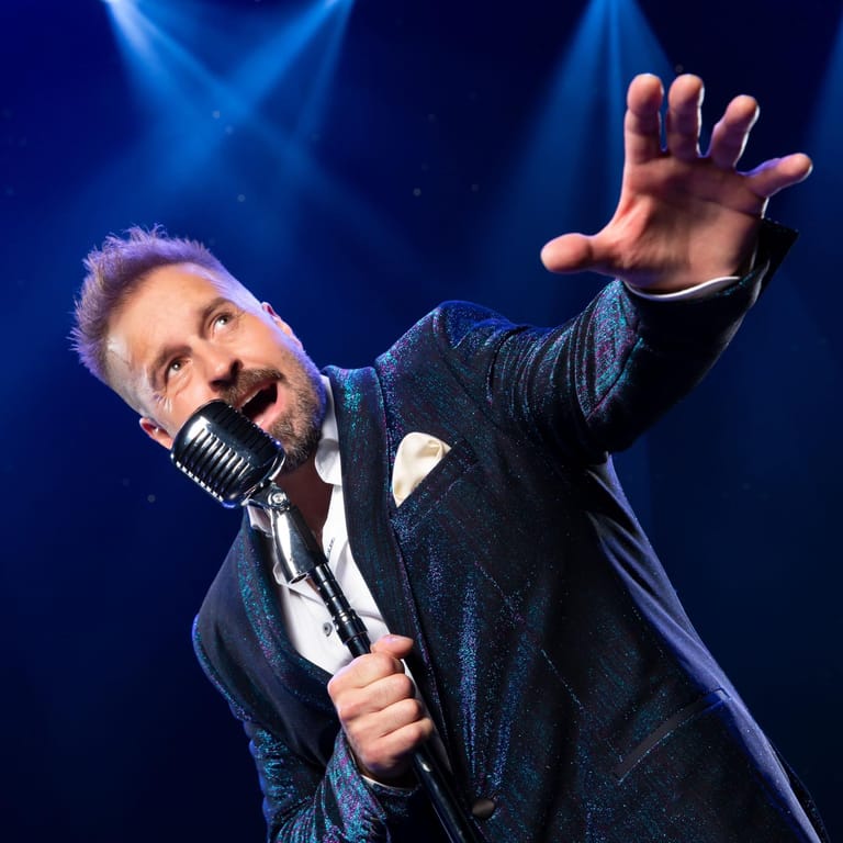 Performer in a dark shimmer jacket holds a microphone with one arm extended towards the audience under blue stage lighting.
