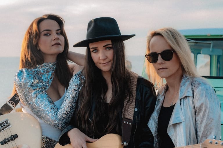 Three women pose together outdoors with a vintage caravan, wearing casual style with hats and sunglasses.