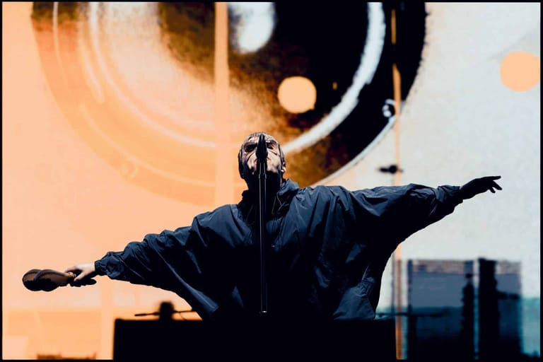 Performer in black jacket with arms outstretched against warm-toned stage lighting and geometric backdrop.