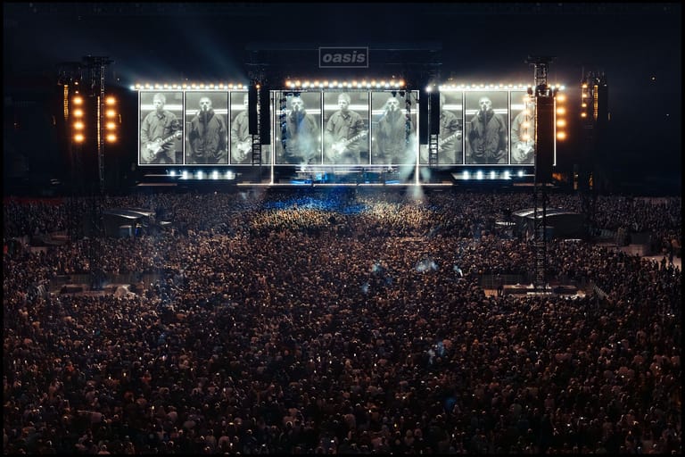 Massive crowd fills an arena beneath illuminated stage screens displaying band members during a live concert tour.
