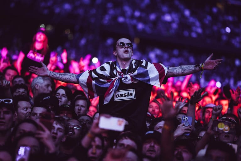 Performer in striped vest with arms spread wide, surrounded by crowd under purple stage lighting during a concert tour.