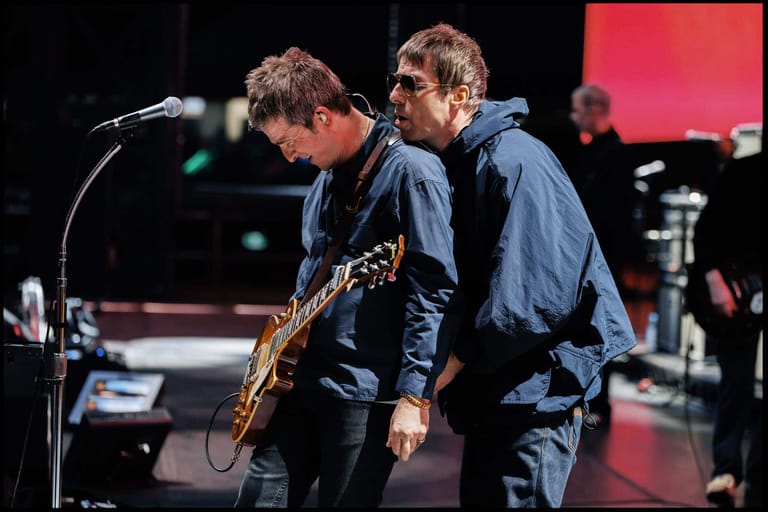 Two musicians perform on stage, one singing at a microphone and the other playing saxophone, both wearing dark jackets.