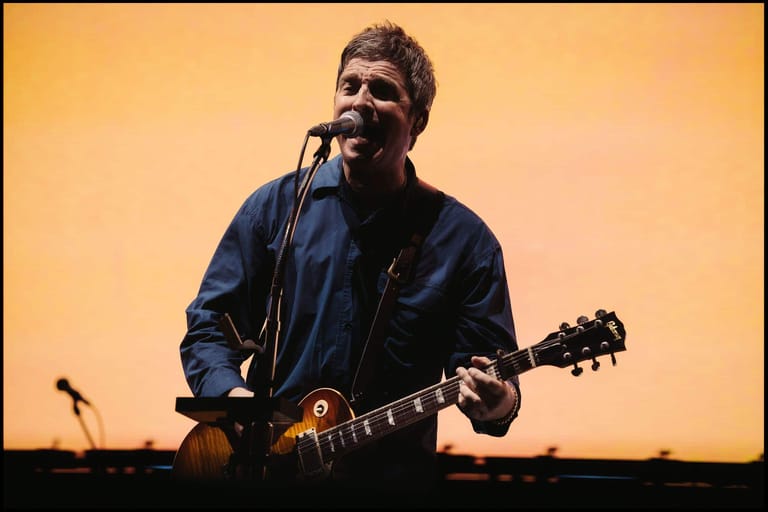 Guitarist and vocalist performing with electric guitar against a warm golden stage backdrop.
