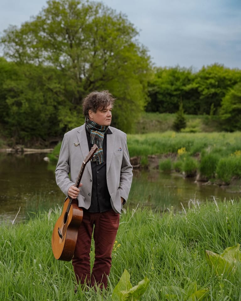 Musician holding an acoustic guitar stands in a grassy field beside a river, surrounded by green trees.