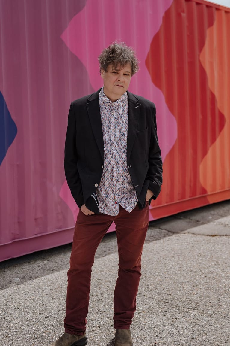 Man in black blazer and burgundy trousers standing against a vibrant pink and red geometric mural backdrop.