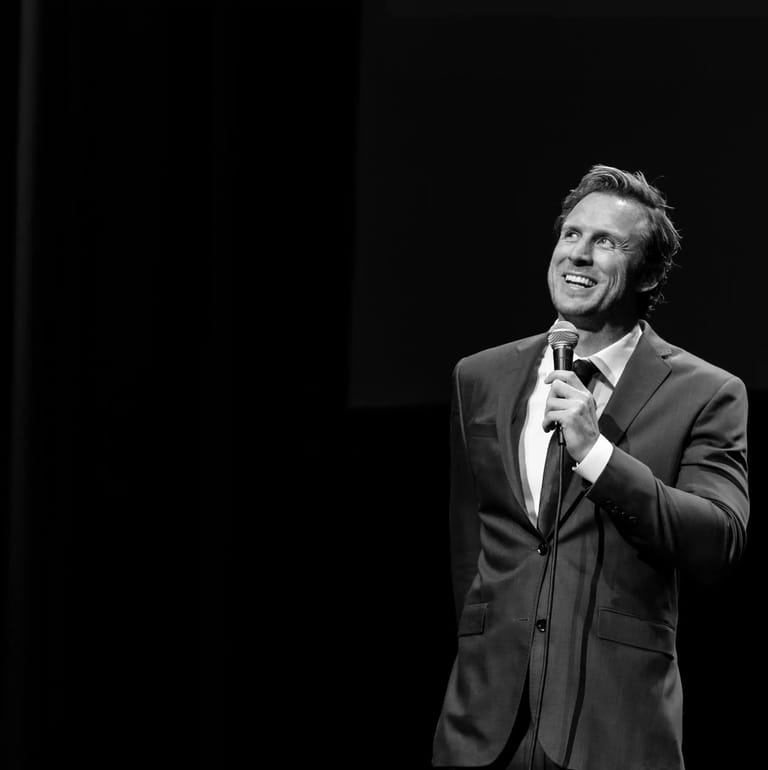 Man in dark suit and tie holding microphone, smiling against black stage background.