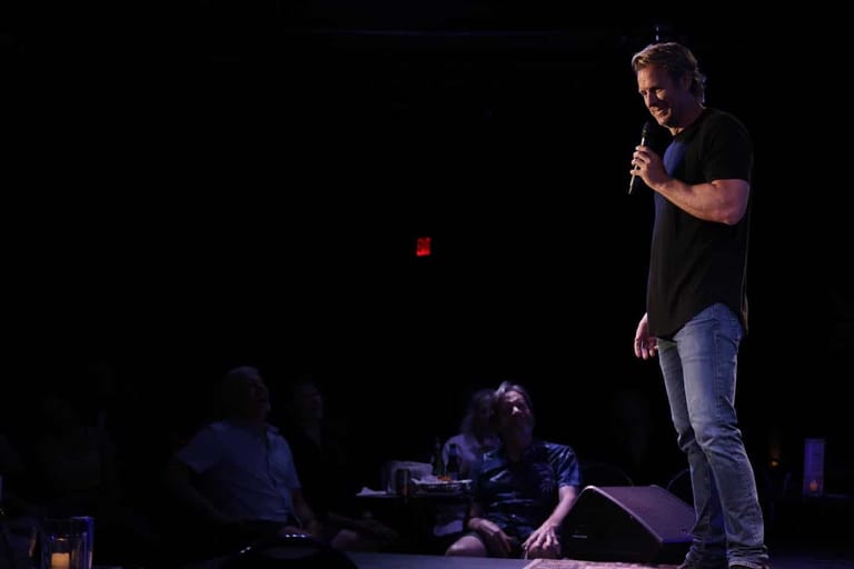 Man in dark shirt and jeans speaking into microphone on dimly lit stage with seated audience members.