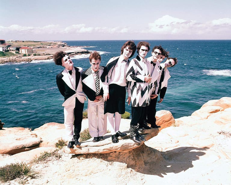 Five performers in formal attire pose on a rocky clifftop overlooking turquoise ocean and coastal landscape.
