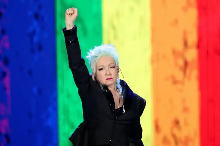 Performer with white hair in black blazer raises fist against rainbow-coloured stage lighting.