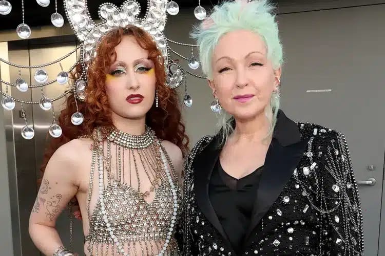Two performers in glamorous stage outfits with dramatic makeup and styled hair stand together against a sparkling backdrop.