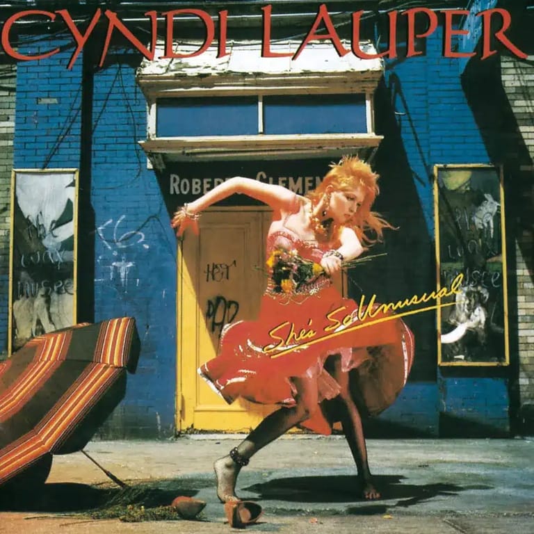 Woman with red-blonde curly hair in a beige corset and red skirt poses dynamically against a blue building with yellow door.