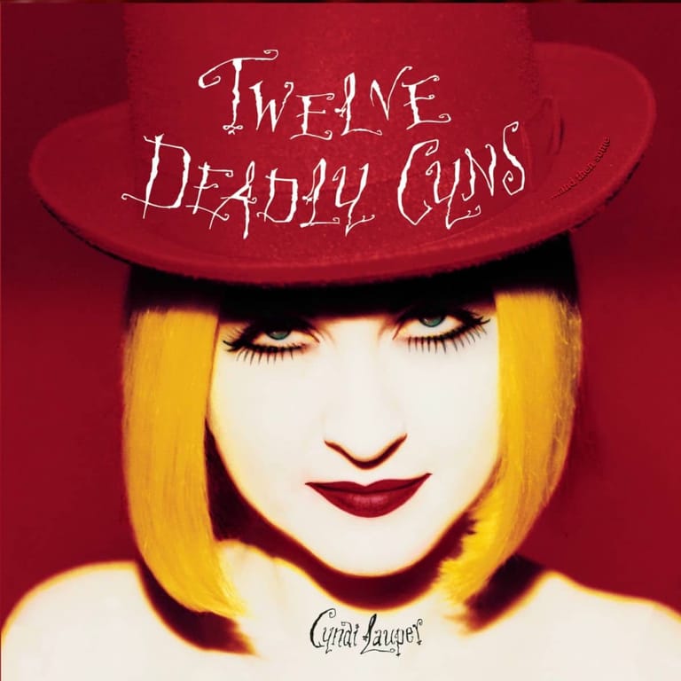 Person with yellow bob hair and dark lipstick wearing a burgundy hat against a deep red background.