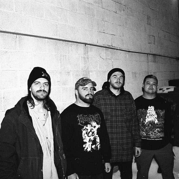 Four band members stand against a concrete wall in black and white, wearing casual attire including beanies and graphic band shirts.